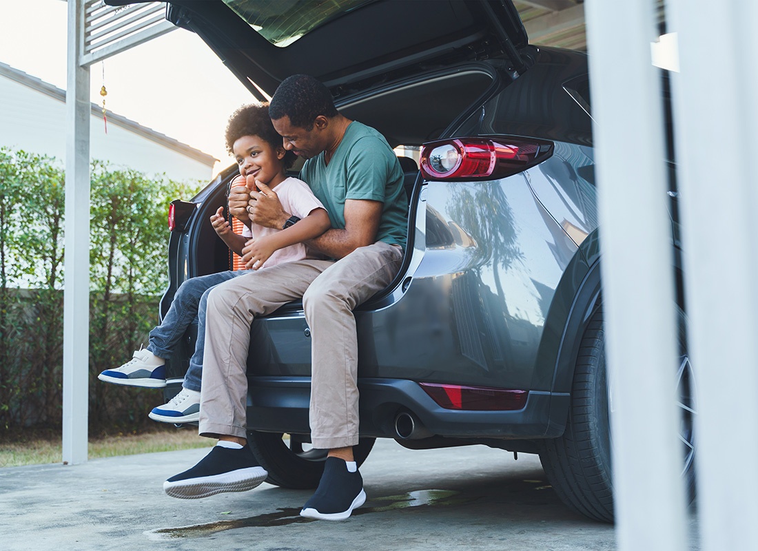 Insurance Solutions - Portrait of a Father and Son Sitting in the Back Trunk of a Car Parked in the Driveway Under a Carport Getting Ready to Go on a Road Trip